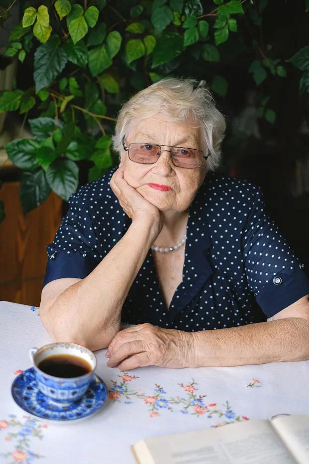 An older woman at her kitchen table with a cup of tea and a book, surrounded by houseplants.