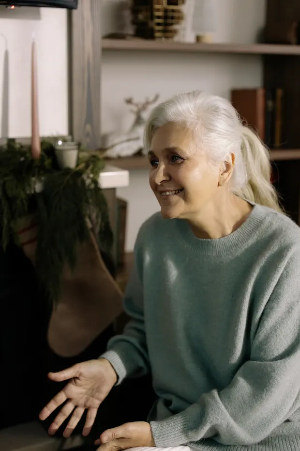 A smiling older woman at home in a warm sweater, surrounded by familiar comforts.