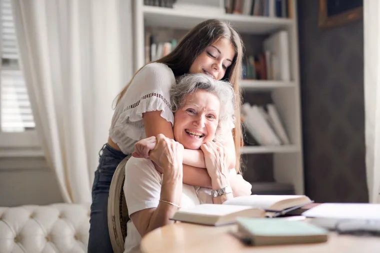 A granddaughter leaning in to hug her smiling grandmother at home.