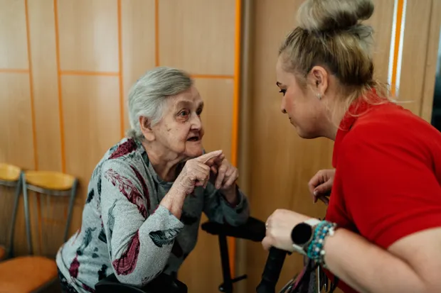 A caregiver crouching down to talk with an older woman, listening closely to what she says.