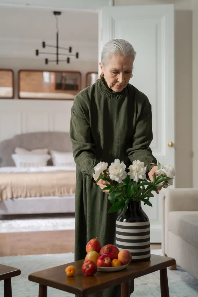 An older woman arranging fresh flowers in a vase in her own living room.