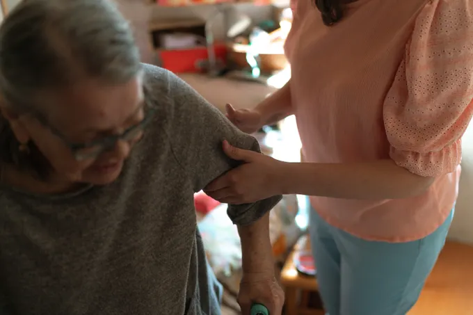 A caregiver steadying an older woman’s arm as she moves through her living room.